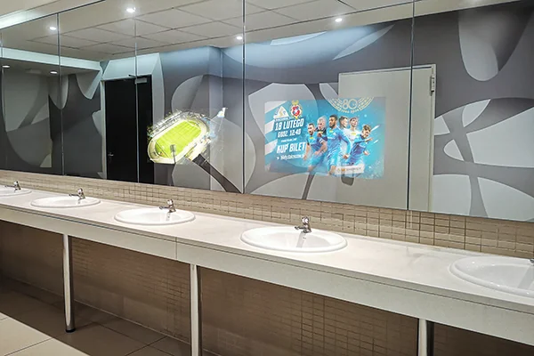 Zdjęcie łazienki w galerii handlowej z wyświetlanymi reklamami na lustrach ponad umywalkami.