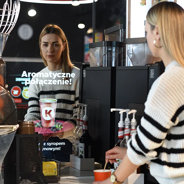Photo at a coffee machine at a gas station. On the wall next to the coffee machine is a mirror displaying an advertisement, with a woman standing in front of the coffee machine looking at the ad.