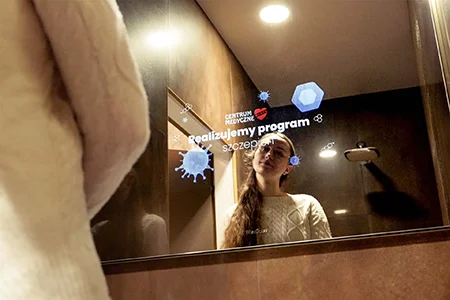 Photo of a woman in a brown bathroom. The woman is looking at her reflection in the mirror, which displays an advertisement for the MEDYK Medical Center.