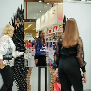 Zdjęcie lustra na czarnym wysokim stojaku. Dwie kobiety przyglądają się lustrze. Na lustrze wyświetla się reklama WiseGlass. W odbiciu lustra widać wnętrze galerii handlowej.