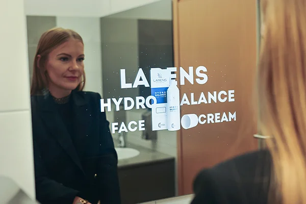 Zdjęcie w łazience. Przed lustrem stoi kobieta, patrzy się na reklamę Larens wyświetlaną na lustrze przed nią.
