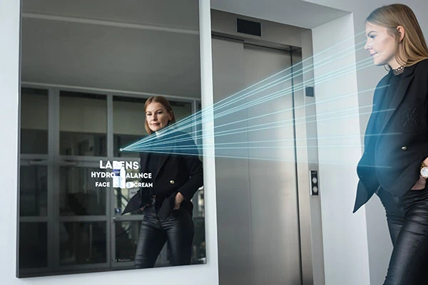 Kobieta mija lustro reklamowe obok windy. Wyimaginowane promienie z lustra skanują kobietę.