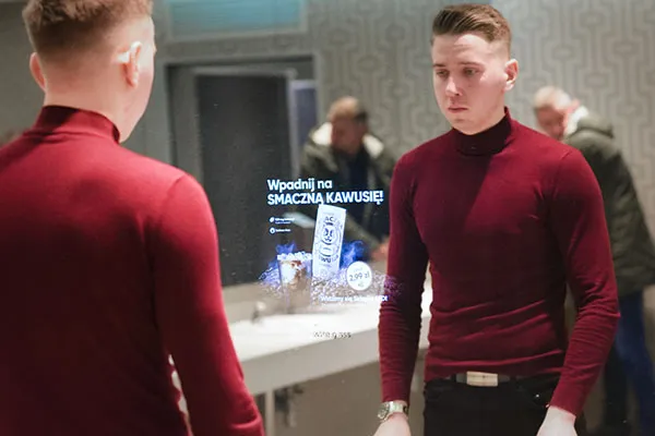Mężczyzna w łazience stoi przed lustrem reklamowym,=. Na lustrze wyświetla się reklama kawy.